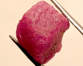 May include: A rough, pink ruby gemstone held by tweezers. The ruby has a natural, unpolished finish.