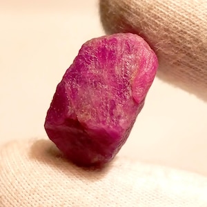 Puede incluir: Una gema de rubí en bruto sin cortar. El rubí es de un color rojo rosado profundo y vibrante con una superficie rugosa y texturizada. La gema se sostiene sobre un fondo blanco.