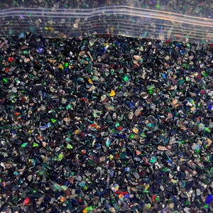 May include: A collection of small, crushed black opal fragments contained within a clear plastic resealable bag. The tiny, irregular stone pieces feature a dark base color with vibrant, iridescent flashes of green, red, blue, and yellow light.