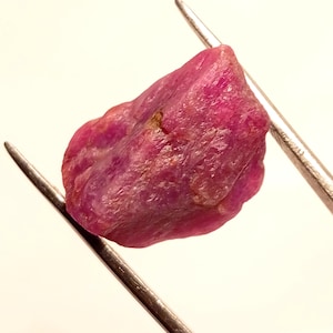 Puede incluir: Una piedra preciosa de rubí rosa en bruto con una forma natural irregular. El rubí se mantiene en su lugar por dos pinzas metálicas.
