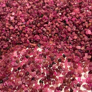 May include: A large collection of small, raw, unpolished gemstone fragments in various shades of deep pink, magenta, and dark red. The stones are irregular in shape and size, featuring a translucent, crystalline texture with scattered dark inclusions.