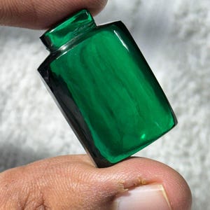 May include: A rectangular, emerald green glass bottle with a small, square neck. The bottle has a glossy finish and a dark, almost black, base. The bottle is held between fingers.