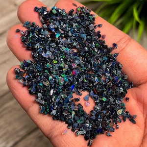 May include: A collection of small, irregular black opal chips featuring vibrant flashes of iridescent color including green, blue, and red. These tiny, dark, polished gemstone fragments exhibit a high-gloss finish and are scattered across the palm.