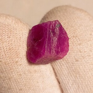 May include: A rough, uncut ruby gemstone. The ruby is a deep, vibrant pinkish-red colour, with a rough, textured surface. The gemstone is held against a white fabric background.