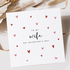 May include: A white Valentine's Day card with red and pink hearts scattered across the surface. The card reads "To my wife on Valentine's Day" in a handwritten style font. The card is square and sits on a white surface.