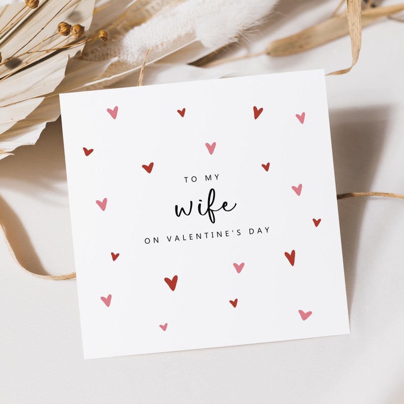 May include: A white Valentine's Day card with red and pink hearts scattered across the surface. The card reads "To my wife on Valentine's Day" in a handwritten style font. The card is square and sits on a white surface.