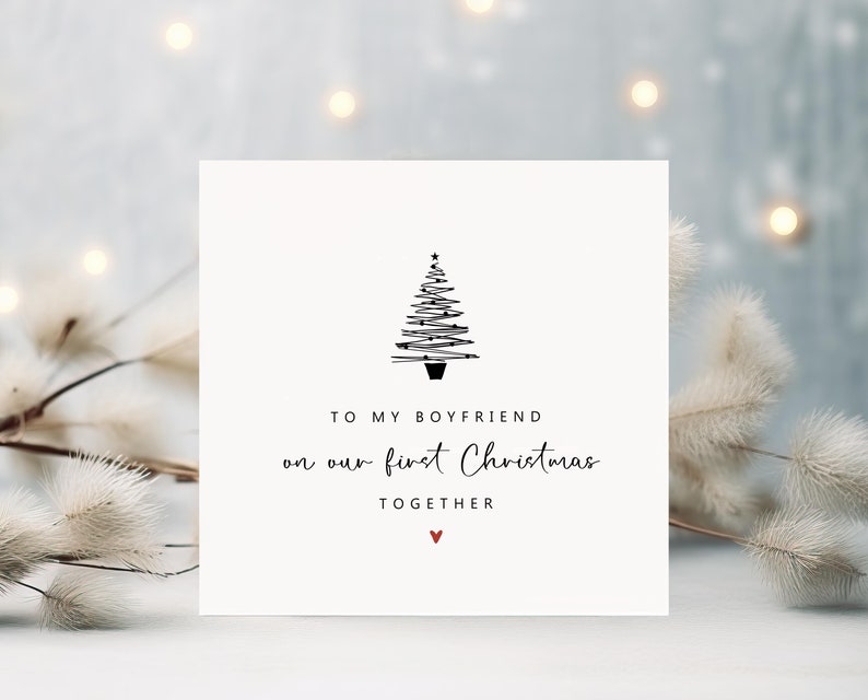 K&ouml;nnte beinhalten: Quadratische Weihnachtskarte mit minimalistischem Design. Die Karte zeigt eine schwarze Strichzeichnung eines Weihnachtsbaums und den Text "TO MY BOYFRIEND on our first Christmas TOGETHER" mit einem kleinen roten Herz.
