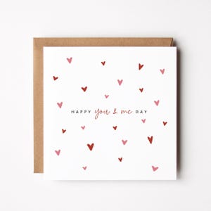 May include: A square greeting card with a brown envelope. The white card features scattered red and pink hearts. The words "HAPPY you & me DAY" are printed in a delicate font, perfect for Valentine's Day or anniversaries.