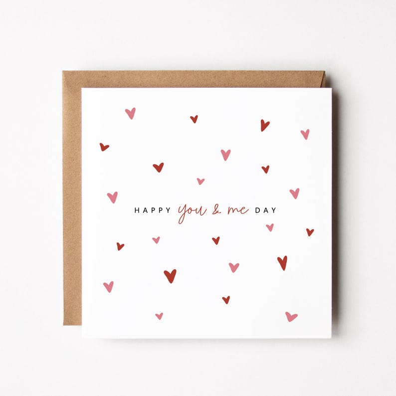 May include: A square greeting card with a brown envelope. The white card features scattered red and pink hearts. The words "HAPPY you & me DAY" are printed in a delicate font, perfect for Valentine's Day or anniversaries.