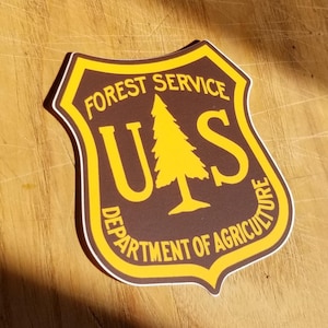 May include: A brown and yellow shield-shaped sticker with the text "FOREST SERVICE" and "DEPARTMENT OF AGRICULTURE" surrounding a stylized tree with the letters "US" in the center.