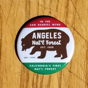 May include: A red, white, and green button with a brown bear silhouette. The button reads "In the San Gabriel Mtns Angeles Nat'l Forest Est. 1908 ANF USFS California's First Nat'l Forest".