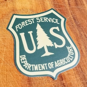May include: A green and beige shield-shaped sticker with the text "FOREST SERVICE" and "U.S. DEPARTMENT OF AGRICULTURE" surrounding a stylized evergreen tree.