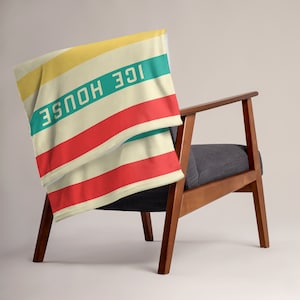May include: A folded striped throw blanket with the words "ICE HOUSE" in green and yellow lettering. The blanket is draped over a wooden armchair with a gray cushion.