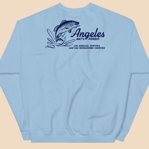 May include: Light blue sweatshirt with a graphic of a fish jumping out of the water. The text "Angeles Nat'l Forest" is printed on the sweatshirt, along with the words "Los Angeles, Ventura and San Bernardino Counties".