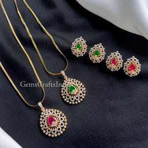 Può includere: Set di gioielli color oro con due collane con pendenti e due paia di orecchini. I pendenti e gli orecchini hanno un design floreale con pietre preziose verdi e rosse, circondate da piccole pietre trasparenti. Il testo "GemsCraftsIndia" è visibile.