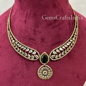 May include: A gold necklace with a teardrop-shaped green stone pendant and a round pendant with white stones. The necklace is on a red velvet background. GemsCraftsIndia is written in white text on the background.
