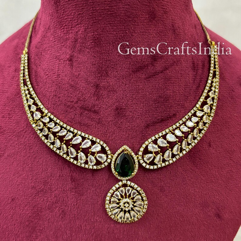 May include: A gold necklace with a teardrop-shaped green stone pendant and a round pendant with white stones. The necklace is on a red velvet background. GemsCraftsIndia is written in white text on the background.