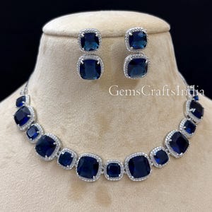 Puede incluir: Un collar y pendientes de plata con grandes piedras azules cuadradas rodeadas de piedras transparentes m&aacute;s peque&ntilde;as. La joyer&iacute;a se muestra en un maniqu&iacute;.