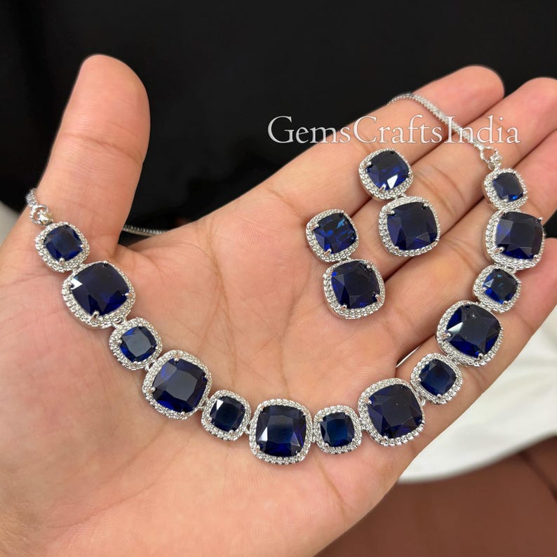 Puede incluir: Un collar y pendientes de plata con grandes piedras cuadradas de color azul oscuro rodeadas de piedras m&aacute;s peque&ntilde;as y transparentes. El texto "GemsCraftsIndia" es visible en la imagen.