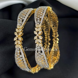 Puede incluir: Dos brazaletes dorados con intrincados diseños florales y geométricos. Los brazaletes están incrustados con brillantes piedras transparentes, creando un efecto deslumbrante. El texto "GemsCraftsIndia" es visible. Las joyas se exhiben sobre un fondo negro.