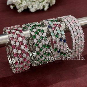 Op de afbeelding: Vier zilveren armbanden met rijen van heldere zirkonia-stenen en gekleurde edelstenen. De armbanden zijn gestapeld op een zilveren oppervlak. De edelstenen zijn roze, groen, blauw en lichtblauw.