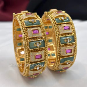 Può includere: Due bracciali rigidi color oro con inserti rettangolari alternati in smalto verde salvia e crema con accenti rosa. Ogni segmento è bordato da dettagli in metallo dorato a perline e include piccole pietre chiare incastonate nei pannelli verdi.