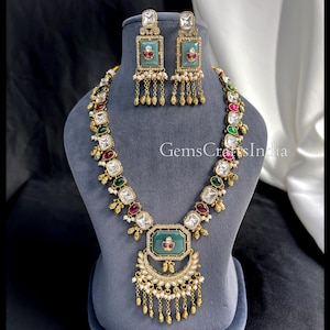 Könnte beinhalten: Ein verziertes, goldfarbenes Halsketten- und Ohrring-Set. Die Halskette hat einen großen, grünen, rechteckigen Anhänger mit floralem Design, umgeben von klaren und farbigen Edelsteinen. Die Ohrringe haben ein ähnliches Design. Das Set wird auf einer grauen Büste präsentiert.
