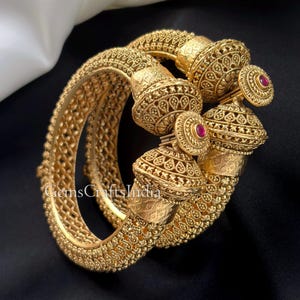 May include: Ornate gold-colored bangles with intricate patterns and a textured finish. The bangles feature decorative bell-shaped elements with small red gemstones. The design includes detailed filigree and embossed textures, showcasing a vintage aesthetic.