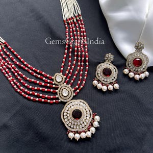 May include: A necklace and earring set with a red and white bead design. The necklace has multiple strands and a large pendant with a central red stone. The earrings and pendant are adorned with clear stones and white pearl drops, creating a luxurious look.