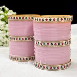 May include: Two pink bangles with gold and white accents. The bangles are decorated with a pattern of small circles in white, green, and red.