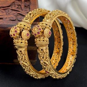 May include: Two gold-toned bangles featuring intricate floral engravings and granulated metal textures. Each bangle is accented with small, circular clusters of deep pink stones set into the metalwork, creating a detailed and ornate appearance.