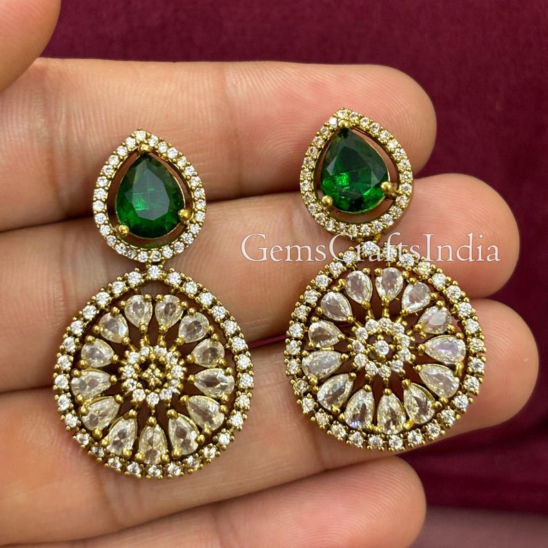 May include: Gold tone drop earrings with a round design featuring clear stones and a green pear-shaped stone at the top.