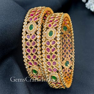 May include: Two gold-colored bangles with intricate detailing. Each bangle is adorned with red and green gemstones, and small clear stones. The text "GemsCraftsIndia" is visible on the bottom of the image. These bangles are a type of jewelry.