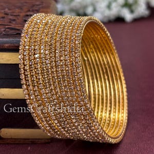 Puede incluir: Brazaletes dorados adornados con pequeñas gemas brillantes. Los brazaletes están apilados, creando una pulsera gruesa y ornamentada. El texto "GemsCraftsIndia" es visible. Las joyas se exhiben sobre una superficie de color rojo oscuro.