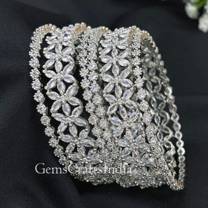 May include: A set of silver-toned bangles with a floral design. Each bangle is adorned with clear, sparkling stones arranged in a flower pattern. The bangles are displayed on a black surface, showcasing their intricate details.