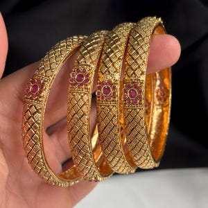 May include: Four gold bangles with a textured, diamond-patterned design. Each bangle features a small, round, red gemstone flower. The bangles are held in a hand, showcasing their intricate details and warm, golden hue.