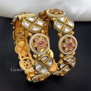 May include: Two gold-toned hinged bangles featuring alternating circular floral motifs with central pink stones and white bead borders, interspersed with geometric segments containing clear, flat-cut stones. The metalwork is detailed and ornate.