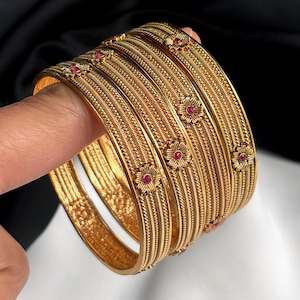 Peut inclure: Ensemble de bracelets dorés avec des détails texturés complexes. Chaque bracelet est orné de petites fleurs rouges. Les bracelets sont empilés, mettant en valeur leur design. Les bijoux sont présentés sur un fond sombre.