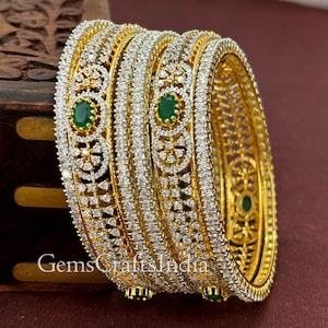 Könnte beinhalten: Satz von vier goldenen Armreifen mit aufwendigen Designs aus weißen Steinen und grünen Steinakzenten. Die Armreifen sind übereinander gestapelt.
