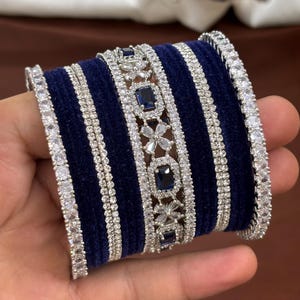 Op de afbeelding: Een stapel marineblauwe fluwelen armbanden met zilveren en heldere strass-accenten. Sommige armbanden hebben rechthoekige en bloemvormige donkerblauwe edelstenen. De armbanden worden in een hand gehouden.