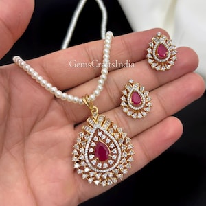 May include: A jewellery set featuring a pearl necklace with a gold-toned pendant and matching earrings. The pendant and earrings are teardrop-shaped, set with a central red stone surrounded by clear white stones. The text GemsCraftsIndia is visible.