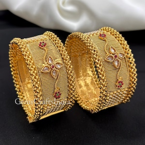 Puede incluir: Dos brazaletes dorados con una banda central de malla texturizada, bordeada por intrincados bordes trenzados. Cada brazalete muestra un motivo floral central con piedras claras y pequeños acentos rojos y verdes. El texto GemsCraftsIndia es visible.