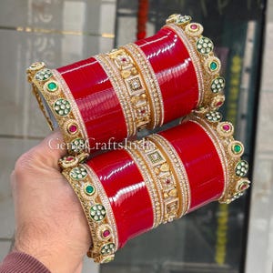 Può includere: Due braccialetti rossi con decorazioni dorate. I braccialetti presentano intricati disegni dorati con pietre preziose incastonate in verde, rosso e bianco. Il testo "GemsCraftsIndia" è visibile nell'immagine.