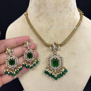 May include: A gold-toned necklace and earring set. The necklace features a pendant with a large emerald-green rectangular stone, surrounded by smaller clear stones. The earrings mirror the pendant design. Both pieces have dangling green beads and small white pearls.
