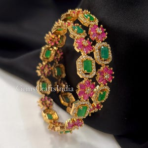 Peut inclure: Bracelet jonc doré orné de pierres précieuses vertes taillées en émeraude, d'accents floraux roses et de cristaux transparents. Le bracelet présente un motif répétitif de pierres vertes et de fleurs roses. La marque "GemsCraftsIndia" est visible.