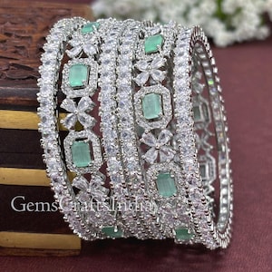 May include: A set of silver-toned bangles with emerald-green rectangular stones and clear, sparkling accents. The bangles feature floral and geometric designs, with the text "GemsCraftsIndia" visible on the bottom left.