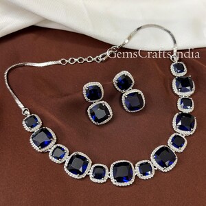 Puede incluir: Un collar y pendientes de plata con grandes piedras azules cuadradas rodeadas de piedras transparentes m&aacute;s peque&ntilde;as. El collar tiene un dise&ntilde;o de cadena.