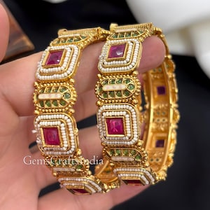 Peut inclure: Deux bracelets rigides dorés ornés de pierres magenta de forme carrée entourées de rangées de petites perles blanches. La structure métallique présente des accents verts et des détails en émail blanc. Le texte GemsCraftsIndia est visible.
