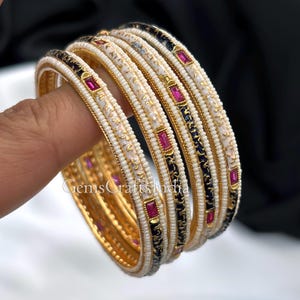 May include: A collection of gold bangles with white and black enamel patterns. Each bangle is adorned with tiny, white beads and rectangular, red gemstones. The bangles are stacked, highlighting their detailed design and craftsmanship.