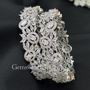 Puede incluir: Dos pulseras plateadas con intrincados adornos florales y cristales transparentes en forma ovalada. Las pulseras están apiladas, mostrando el diseño detallado. El texto "GemsCraftsIndia" es visible. El fondo es negro.